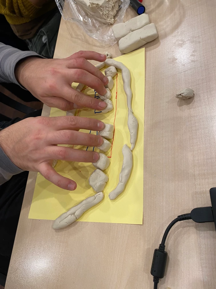 Knetmasserollen umranden einen gezeichneten Entwurf. Knetmasse-Würfel stellen fühlbare Tasten dar, auf denen die Finger von zwei Händen liegen.