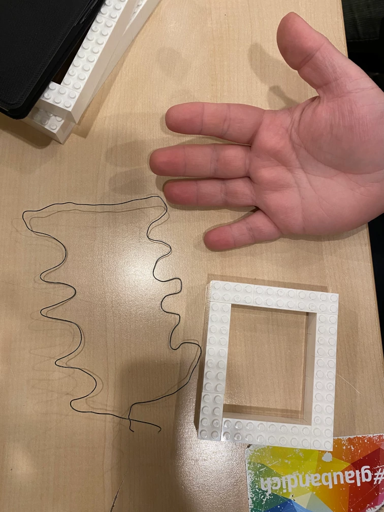 Draht in Form von Handgriffen mit Vertiefungen für jeden Finger. Rechteck aus Klemmbausteinen. Hand im Bild zum Größenvergleich.