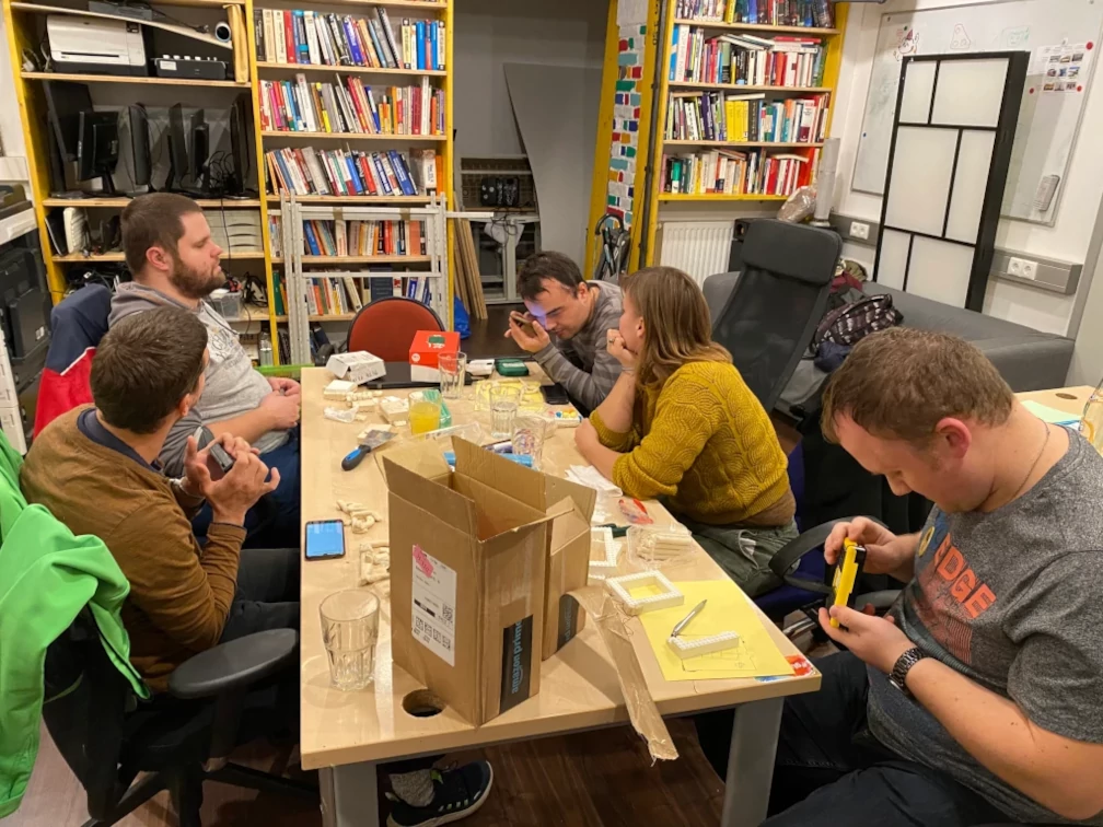 Fünf Menschen sitzen an einem Tisch. Sie beschäftigen sich mit Tastaturen und Smartphones in ihren Händen oder verfolgen die Arbeit der Person nebenan. Auf dem Tisch stehen Trinkgläser, geöffnete Kartons, Smartphones, Tastaturen, Klemmbausteine, Knetmasse und noch mehr Arbeitsmaterial. In den Regalen im Hintergrund stehen geschlichtete Bildschirme, Tastaturen, Drucker und viele Bücher. An der rechten Wand steht ein Sofa, eine Wandteiler und ein Whiteboard direkt auf der Wand.