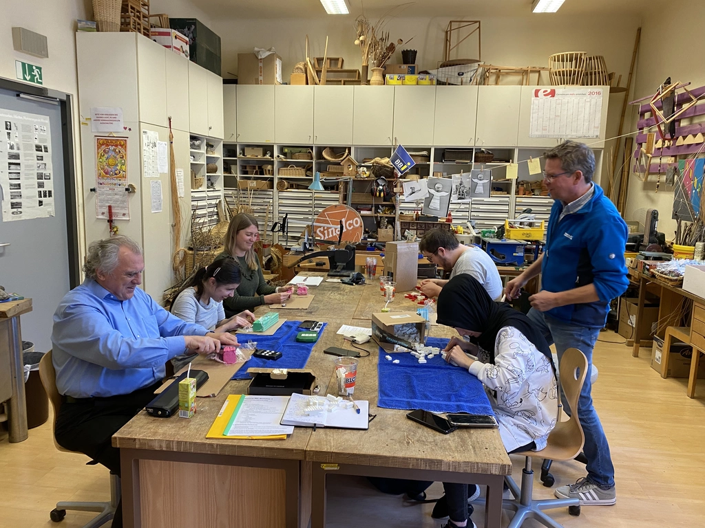 Fünf Menschen sitzen gemeinsam an einem Tisch und eine Person steht neben dem Tisch in einer Werkstatt. Die sitzenden Menschen werken mit Knetmasse und mit Klemmbausteinen. Zwei blaue Handtücher sind auf dem Tisch ausgebreitet, außerdem liegen auf dem Tisch Knetmasse, Klemmbausteine, Unterlagen, Smartphones, Tastaturen, Trinkbehälter, Schachteln und Werkzeug. Im Hintergrund stehen Kästen und Tische voll Arbeitsmaterial und Werkzeug.