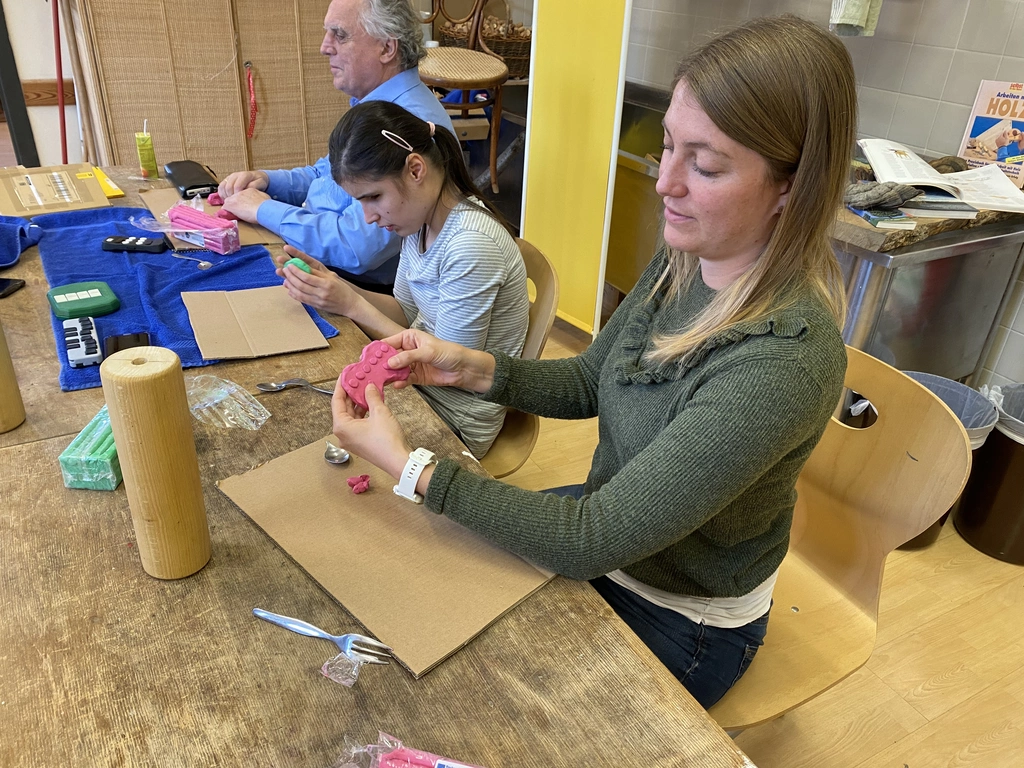 Eine Frau hält einen Prototyp aus Knetmasse in Form einer Spielekonsole vor sich. Am Prototypen sind sechs Knöpfe in einer Reihe über zwei Knöpfen unter ihren Daumen zu erkennen. Sie sitzt mit einer Schülerin und dem Projektleiter auf einer Seite eines Tisches.