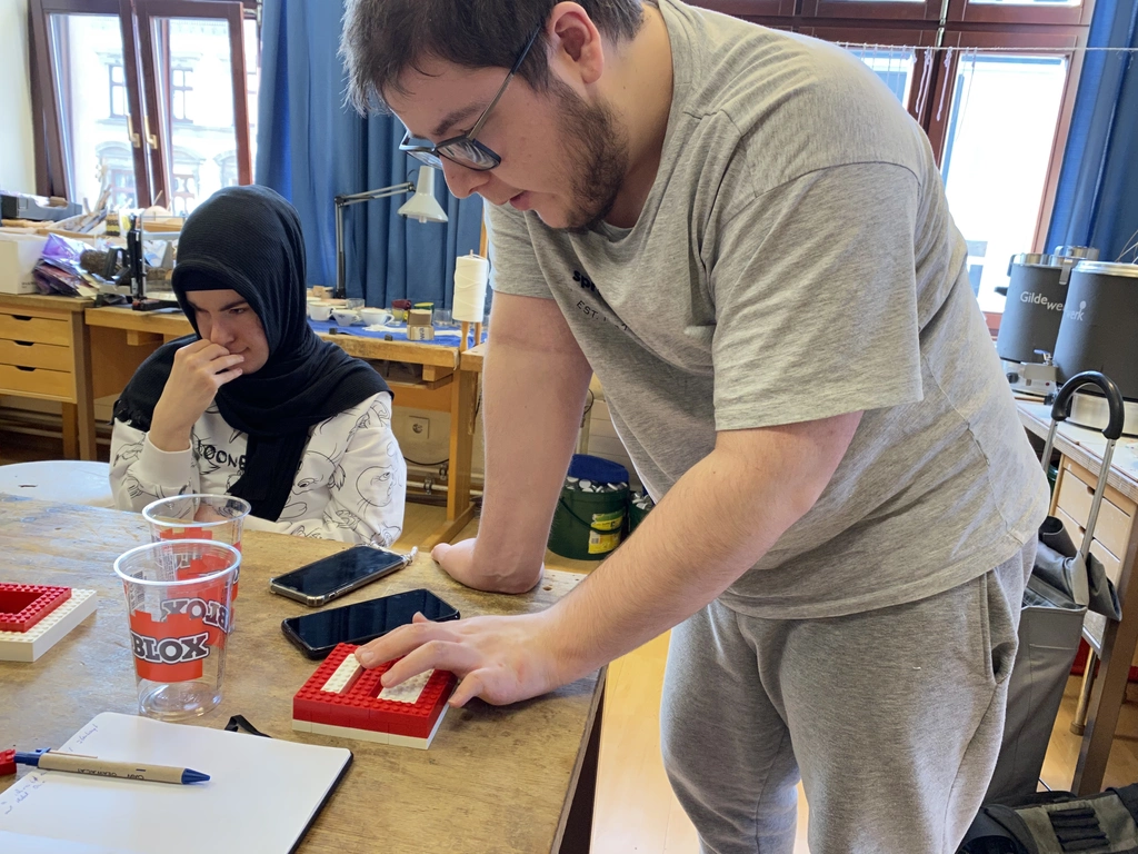 Schüler mit Hand auf einer Braille-Tastatur Prototypen aus Klemmbausteinen auf einem Tisch. Daneben sitzt eine Schülerin. Smartphones, Klemmbausteine, Becher, Stift und Papier liegen am Tisch. Im Hintergrund umrahmen blaue Vorhänge zwei Fenster.