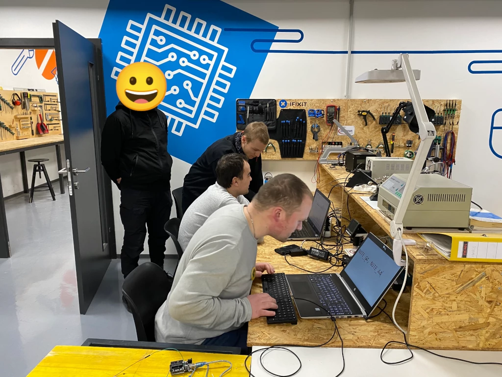 Zwei Personen sitzen vor Laptops. Eine weitere Person beugt sich zum Laptop einer sitzenden Person. Eine unkenntlich gemachte Person steht dahinter. Auf einem Tischaufbau aus Grobspanplatten stehen elektrische Geräte. Im Vordergrund liegt ein über Kabel verbundenes Elektronikbauteil. An der Wand hängen Werkzeuge. Durch eine geöffnete Türe ist weiteres Werkzeug an einer Wand erkennbar. Auf die Wand ist ein Mikrochip in weißer Farbe auf blauen Grund gezeichnet.