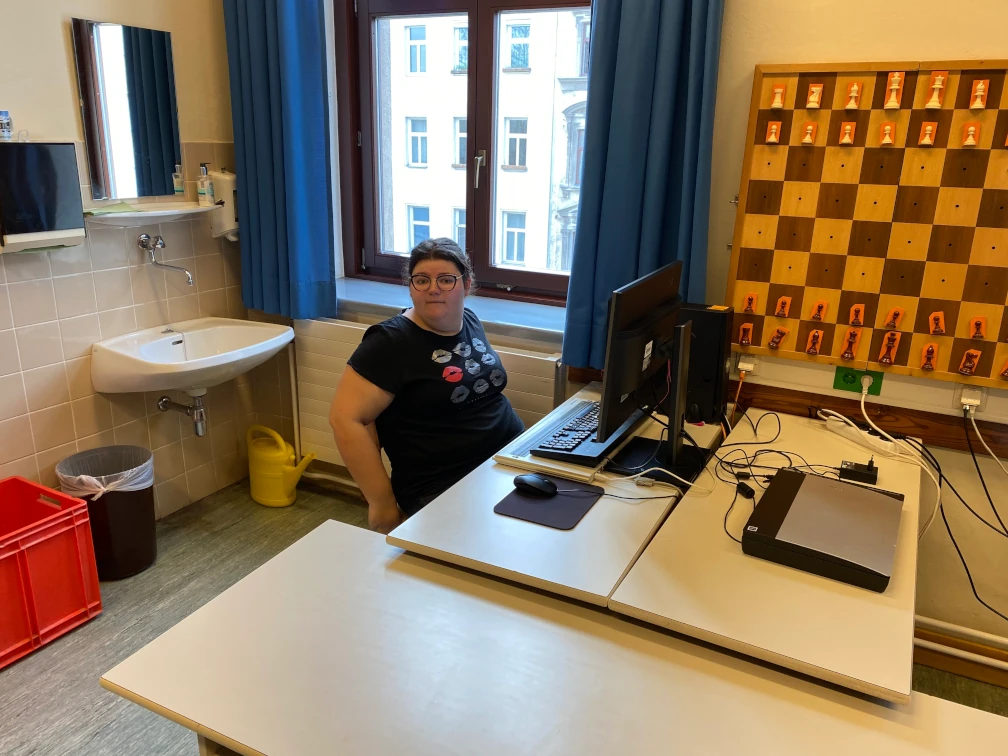 Schülerin mit Brille sitzt an einem Computer-Arbeitsplatz vor einem Monitor, Tastatur, Braillezeile und Maus. Im Hintergrund befindet sich eine Waschgelegenheit, ein Fenster mit Blick auf eine Häuserfront und an der Wand befestigt ist ein Schachbrett mit etwa einem Meter Seitenlänge aus Holz.