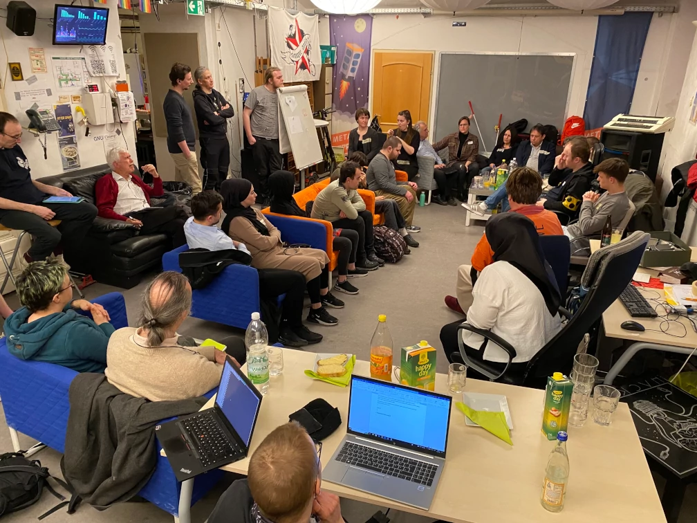 Ca. 24 Personen sitzen in einem Raum auf Sofas und Bürostühlen einander zugewandt, Richtung Raummitte. Auf einem Couch-Tisch und einem Büro-Tisch stehen Getränke und Laptops. Zwei Fahnen mit Abbildungen von Telefonzellen, eine Türe und eine unbeleuchtete graue Anzeige befinden sich an der gegenüberliegenden Wand. Im Hintergrund befindet sich ein Flipchart, ein Telefon und ein Bildschirm mit Balkendiagrammen.