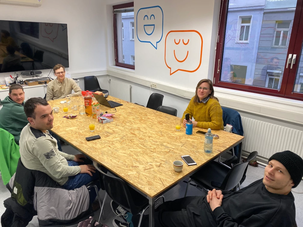 Fünf Menschen sitzen an einem Tisch und lächeln in die Kamera. Auf dem Grobspanplatten-Tisch liegen Smartphones und stehen Getränke sowie ein Laptop. Im Hintergrund befinden sich zwei Fenster, ein großformatiger Bildschirm und Zeichnungen von Sprechblasen mit lächelnden Gesichtern auf der weißen Wand.