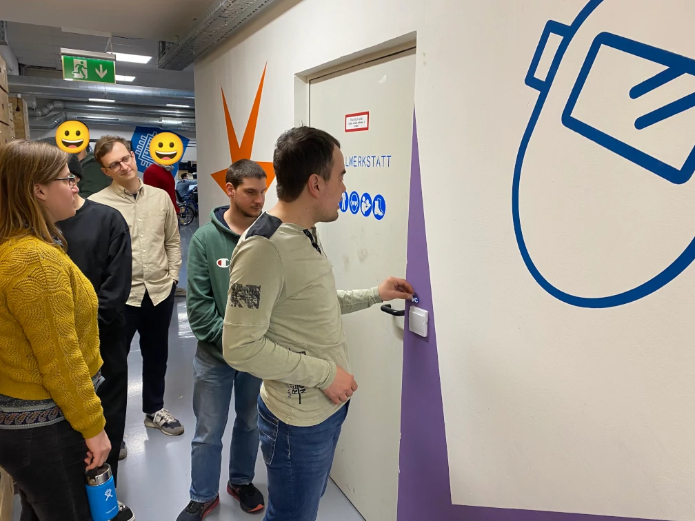 Ein münzgroßes Schild mit den Buchstaben WC wird von einer Person an einen Türrahmen gehalten. Fünf Personen stehen dahinter. Auf der Tür steht Werkstatt und es sind Sicherheitsschilder abgebildet. Auf der weißen Wand rund um die Türe ist ein oranger Stern, eine Schweißermaske in Blau und eine lila Fläche aufgemalt. An der Decke im Gang vor der Türe befindet sich ein Notausgang-Schild.