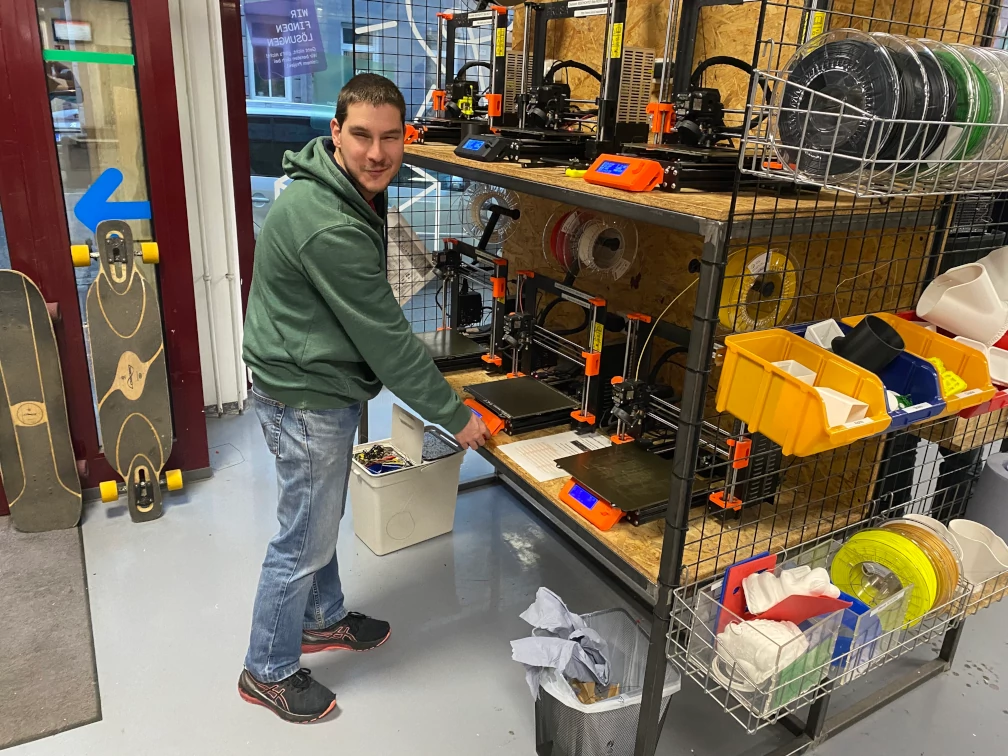 Eine Person lächelt in die Kamera und hat eine Hand am 3D-Druckerbedienfeld. Sechs orange und schwarze 3D-Drucker stehen auf einem Grobspanplatten-Regal. Auf der Seite des Regals befinden sich Filament-Spulen und bereits ausgedruckte Teile in verschiedenen Farben. Im Hintergrund stehen zwei Skateboards an eine Fensterfront gelehnt. Mistkübel stehen unter dem Regal am Boden.
