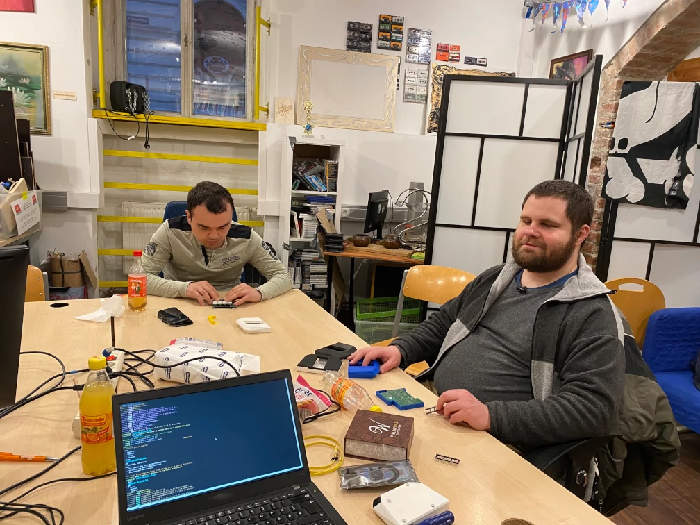 Zwei Menschen sitzen an einem Tisch und arbeiten mit elektronischen Komponenten und Kunststoffgehäusen. Am Tisch steht ein aufgeklappter Laptop, Limonadenflaschen, Schachteln und Papierhandtücher, Steckdosenverteiler und Kabel. In der weißen Wand im Hintergrund ist ein Fenster mit gelben Metallbügeln neben und unter dem Fenster. Raumtrenner verdecken einen weiteren Tisch im Hintergrund zur Hälfte. Sessel stehen vor dem Raumtrenner. Audiokassetten hängen an der Wand.