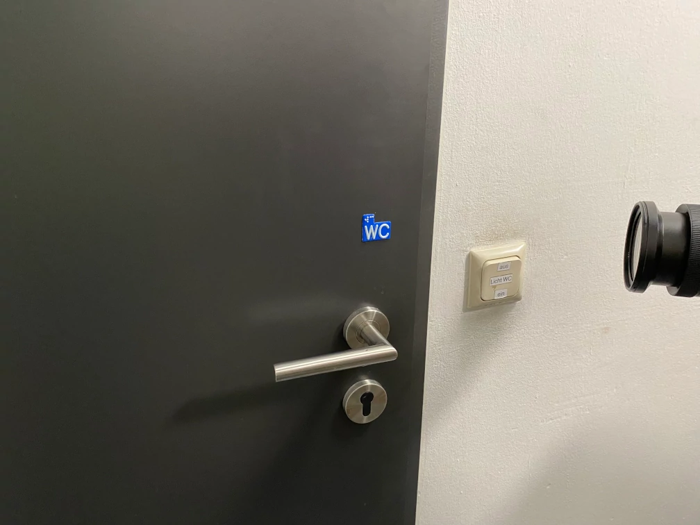 Taktile Beschriftung WC in weißer Schrift auf blauem Grund eine Handbreite über Türgriff einer schwarzen Türe. Das Wort WC steht in Braille-Schrift und in Profil-Schrift auf der taktilen Beschriftung. Eine Kameralinse ist auf das die taktile Beschriftung gerichtet. An der Wand neben der Türe befindet sich ein Lichtschalter mit der Beschriftung "aus; Licht WC; ein".