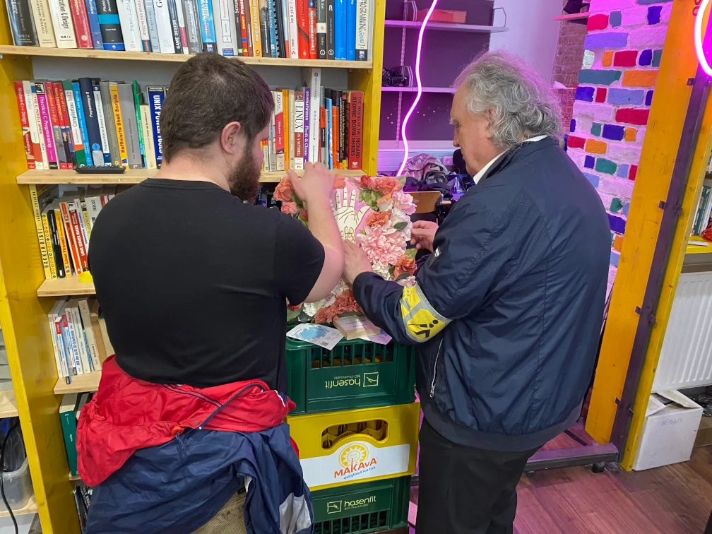 Ein Bild mit Kunstblumen und ausgeschnittenen Händen wird von zwei Menschen begriffen. Auf einem Arm ist eine Blindenschleife erkennbar. Das Bild steht auf umgedrehten Getränkekisten vor einem Bücherregal. Eine Wand mit bunt angemalten Ziegeln führt in einen weiteren Raum.