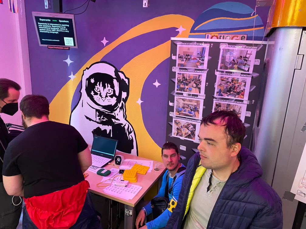 Zwei Menschen stehen vor einem Tisch mit Laptop und ausgedruckten Alphabeten und unterhalten sich. Eine weitere Person scheint in die Kamera zu sehen und eine andere in die Ferne. Auf einem Arm ist eine Blindenschleife erkennbar. Auf einem Rollup befinden sich aufgeklebte Fotos in Klarsichtfolien. Die Wand im Hintergrund ist mit einer Katze im Weltraumanzug auf violett und gelbem Grund bemalt. Eine Ecke einer Telefonzelle ist zu erkennen.