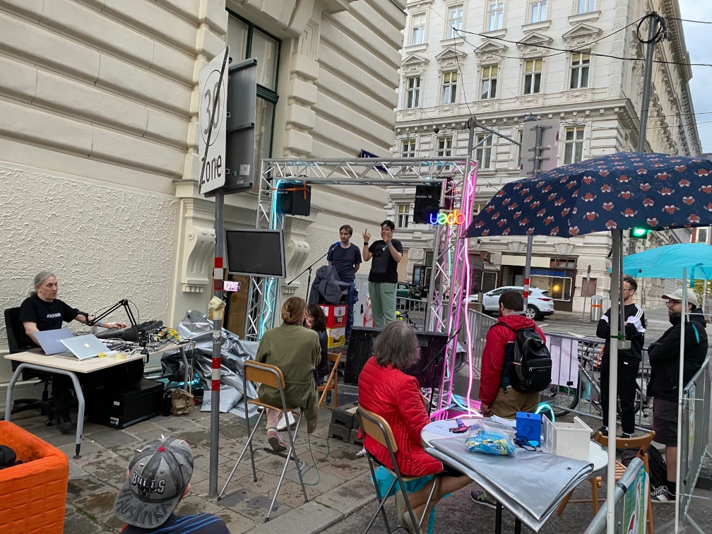 Auf einer mit 4-Punkt-Traversen eingerahmte Bühne stehen zwei Menschen hinter einem Mikrofon, einer gebärdet. Sieben Menschen sitzen oder stehen Richtung Bühne, eine weitere sitzt vor der Bühne, Richtung Publikum. Im Hintergrund befinden sich Häuserfassaden, Absperrgitter und parkende Autos. Ein Verkehrsschild zeigt das Ende der Geschwindigkeitsbegrenzung auf 30 km/h an. Zwei Regenschirme sind wie Sonnenschirme aufgestellt. Auf einem Tisch befindet sich ein Mischpult, ein Mikrofon und zwei Laptops.
