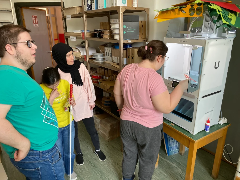 Vier Schüler:innen vor dem 3D-Drucker. Eine Schülerin streckt die Hand zum Ausdruck auf dem Druckbett im 3D-Drucker. Eine Schülerin hält einen Blindenstock. Im Hintergrund ein Regal mit Filamentspulen und eine offene Türe.