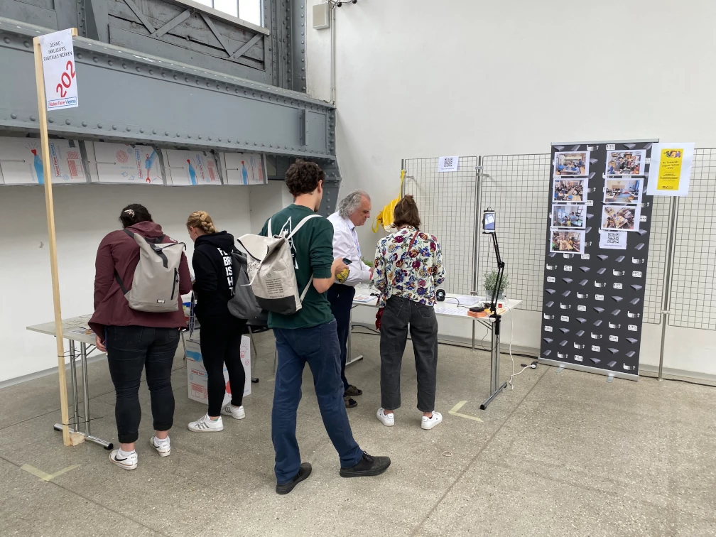 Fünf Personen stehen vor zwei Tischen und betrachten die Gegenstände auf den Tischen oder unterhalten sich. Auf einem Schild steht 'DEFINE - inklusives digitales Werken; 202; Maker Faire Vienna' Ein Smartphone ist mit einem beweglichen Schwenkarm auf einem Tisch befestigt. Fotos, QR-Code und ein Plakat der verrückten Jugend-Aktion VJA sind auf einem Roll-up erkennbar. Ein Drahtgitter, grauer Fußboden, graue Träger eines Stahlskelettbaus, und weiße Wände bilden eine Raumecke im Hintergrund.