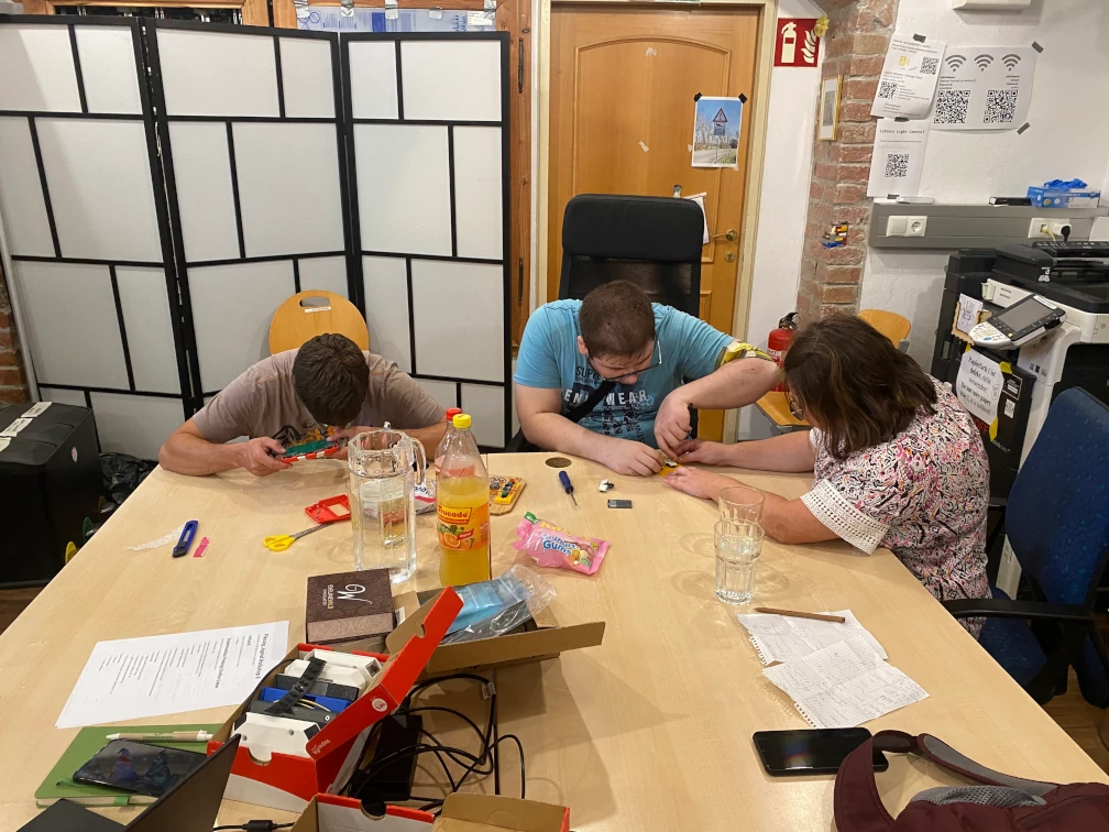 Drei Personen sitzen an einem Tisch und sind in ihre Arbeit vertieft. Die Köpfe sind zu den Gegenständen in ihren Händen geneigt. Zwei arbeiten zusammen an einem Gegenstand am Tisch. Sie sitzen an einem angeräumten Tisch Gläsern, Wasserkrug, Limonade, Messer, Schere, Schachteln, Smartphones und Papieren. Im Hintergrund steht ein Raumteiler und ein Kopierer. In der Wand im Hintergrund befindet sich eine braune Holztüre. An der Wand hängt ein Hinweisschilder zum Feuerlöscher.