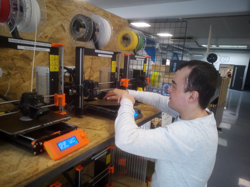 Ein junger Mann wechselt das Federstahlblech vom Druckbett an einem von drei 3-D-Druckern. Die 3-D-Drucker stehen auf einem Regal aus Grobspanplatten. Auf jedem 3-D-Drucker befinden sich zwei Spulen Filament in den Farben rot, weiß, gelb und grün. Auf dem vordersten 3-D-Drucker ist ein runder 3-D-Druck auf dem Druckbett erkennbar.