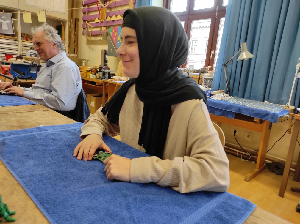 Frau mit schwarzem Kopftuch sitzt an einem Tisch mit Handtuch vor sich ausgebreitet, die Hände liegen auf einer Platine am Handtuch. Weiter hinten sitzt ein Mann mit hellblauem Hemd und grauen Haaren am selben Tisch ebenfalls mit Handtuch vor sich ausgebreitet. Werkbänke, eine Schreibtischlampe und Arbeitsgeräte einer Werkstatt befinden sich hinter den zwei Personen. Tageslicht scheint durch ein Fenster mit blauen Vorhängen in den Raum.