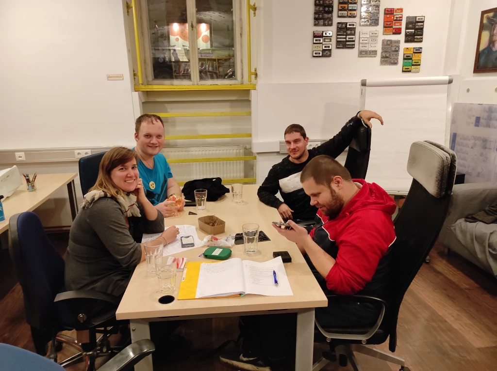 Vier Menschen sitzen an einem Tisch, drei lächeln in die Kamera, einer beschäftigt sich mit einem Smartphone. Auf dem Tisch stehen Trinkgläser, weiters liegen Papiere, Smartphones und kleinere Gegenstände verstreut am Tisch herum. Im Hintergrund ist ein weiterer Tisch, ein Fenster, ein Flipchart und ein Sofa zu erkennen. Auf der weißen Wand im Hintergrund hängen alte Audiokassetten.