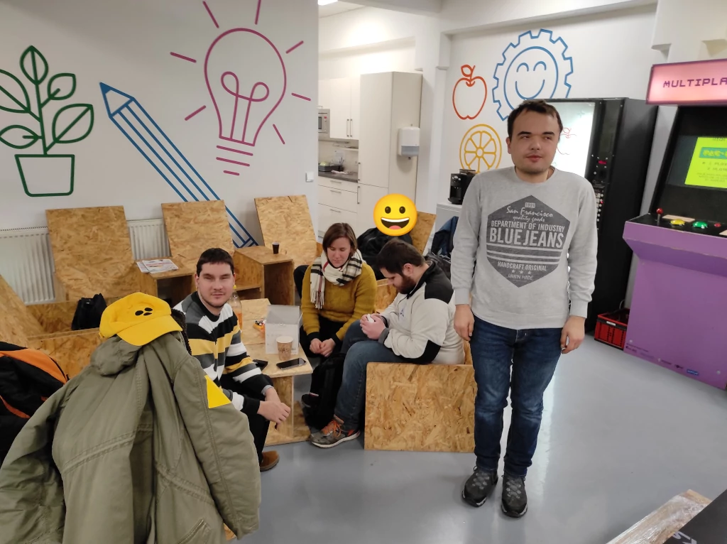Fünf Personen im Bild. Eine Person wurde unkenntlich gemacht, eine Person steht und drei sitzen auf selbstgebauten Sesseln aus Grobspanplatten. Zwei Personen sind auf die Kamera ausgerichtet und zwei weitere sitzen dahinter und unterhalten sich. Im Vordergrund liegt eine gelbe Schirmkappe mit drei schwarzen Punkten zur Kennzeichnung einer Behinderung. Im Hintergrund befinden sich ein Spielautomat, ein Getränkeautomat und eine Küchenzeile. Eine Pflanze, Stift, Glühbirne, Zahnrad mit Gesicht, Apfel und Orange sind in Übergröße auf die weißen Raumwände gezeichnet.