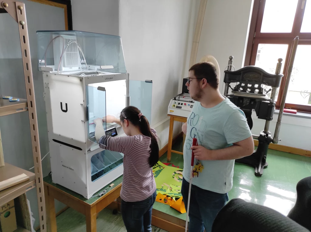 Junge Frau und junger Mann stehen vor 3D-Drucker. Der 3D-Drucker hat die Form eines Schranks mit geöffneten, durchsichtigen Schranktüren. Er ist weiß und von innen beleuchtet. Der 3D-Drucker steht auf einem Tisch und ist ca. einen halben Meter breit und tief und eineinhalb Meter hoch. Die Hände der jungen Frau liegen im 3D-Drucker auf ihrer Kopfhöhe auf einem flachen Ausdruck am Druckbett. Der junge Mann hat einen Blindenstock in der Hand, steht hinter der jungen Frau und ist ebenfalls zum 3D-Drucker ausgerichtet.