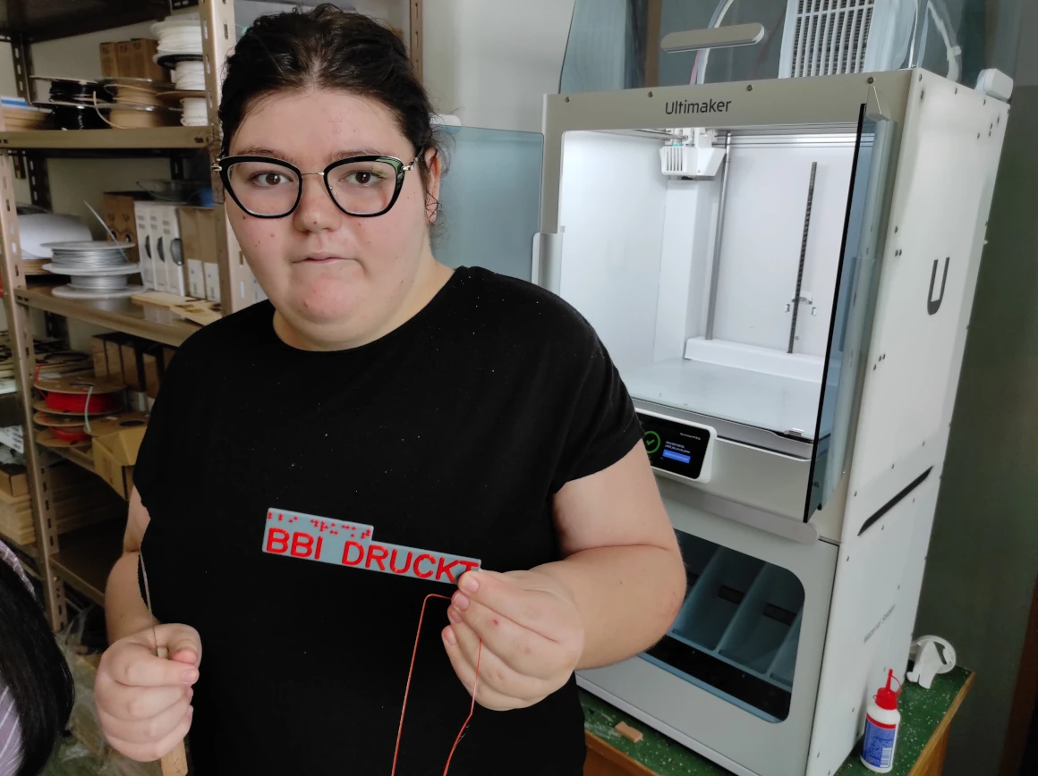 Schülerin, mit Brille, hält 3D-gedrucktes Schild mit der Beschriftung BBI DRUCKT in Profilschrift und Brailleschrift in Rot auf grauem Grund. Sie steht vor einem weißen Ultimaker 3D-Drucker. Im Hintergrund steht ein Regal mit Filamentspulen.