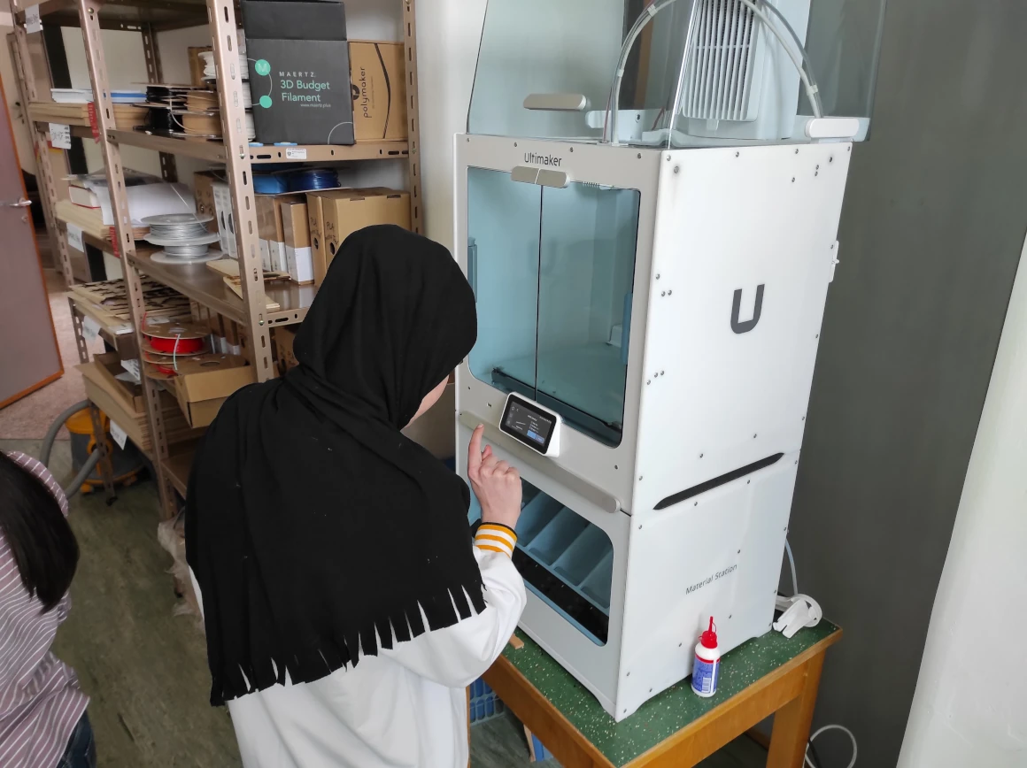 Schülerin, mit Kopftuch, bedient einen Touchscreen eines weißen Ultimaker 3D-Druckers. Im Hintergrund steht ein Regal mit Filamentspulen.