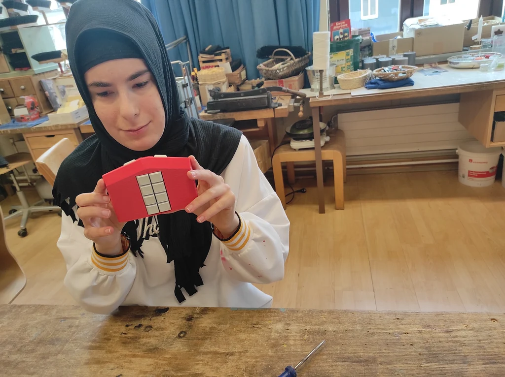Person mit weißer Oberbekleidung und schwarzem Kopftuch hält eine mobile Braille-Tastatur mit weißen Tasten und rotem Gehäuse in beiden Händen vor sich. Im Hintergrund stehen Tisch voll Werkzeugen, Schubladen und Weidenkörben. Durch Fenster sind Hausfassaden erkennbar.