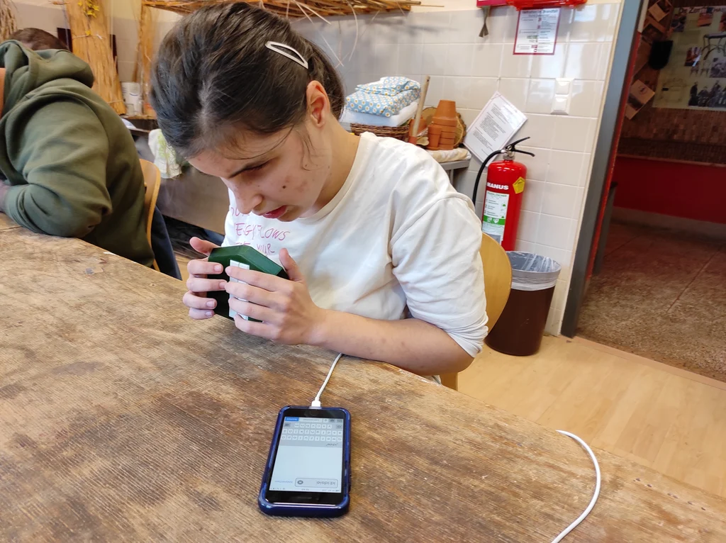 Person mit weißer Oberbekleidung und Haarspange hält eine mobile Braille-Tastatur mit weißen Tasten und grünem Gehäuse in beiden Händen vor sich. Vor ihr liegt ein Smartphone auf dem ein Textfeld und eine virtuelle Tastatur zu sehen sind. Im Hintergrund befindet sich Mistkübel, ein Feuerlöscher an einer gefliesten Wand und ein Durchgang.