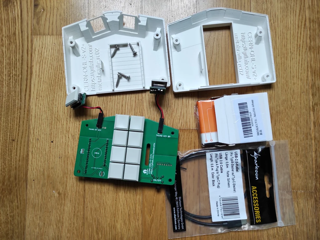 Neun unabhängige Bauteile liegen auf Holz. Die Bauteile sind USB-Kabel, Akku, Metabrailleleiterplatte mit acht Tasten und zwei Tasten hängen an Kabeln an der Platine, zwei weiße Metabraillegehäuseteile und vier Schrauben.
