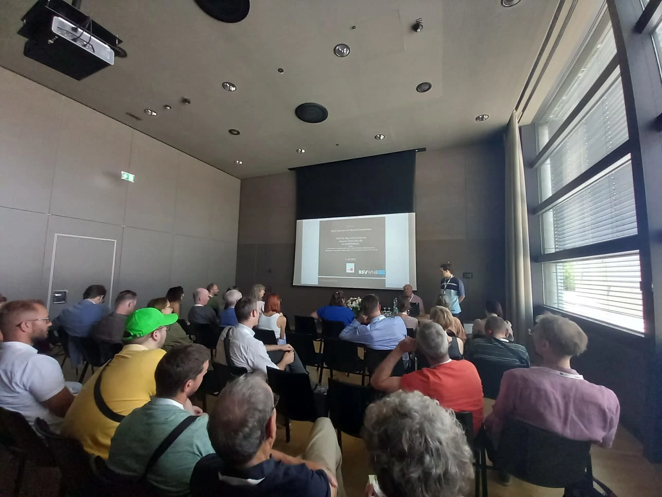 Fast vollbesetzter Saal beim Vortrag über das Projekt Defineben von ca. 36 Sitzplätzen sind unbesetzt. Ein Vortragender sitzt, ein weiterer steht neben einer vom Beamer beleuchteten Leinwand.