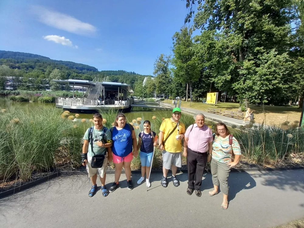 Gruppenbild von sechs Menschen vor dem Uniteich am Johannes Kepler Universitäts Campus. Am grünen Teich ist ein schwimmendes Kaffee zu sehen. Den Hintergrund bilden Wälder und der strahlend blaue Himmel.